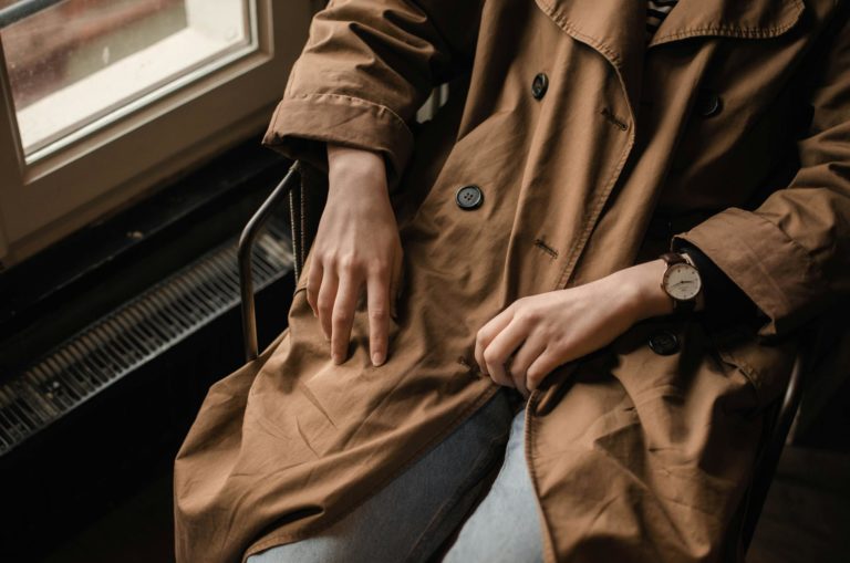 High angle of crop faceless lady in stylish coat and jeans with wristwatch sitting in chair near window in light room in daytime