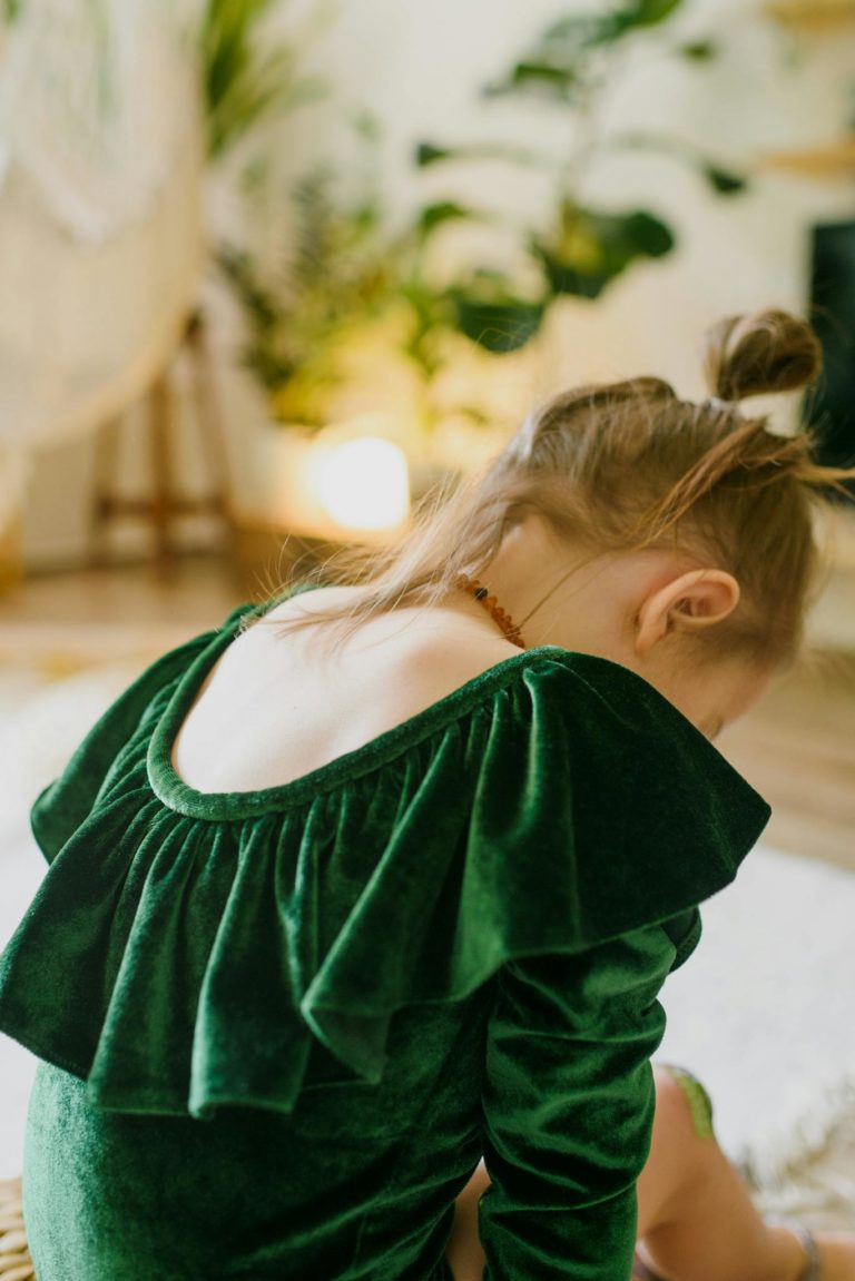 Side view of unrecognizable cute little girl in trendy green velvet dress sitting on floor in stylish room and looking down